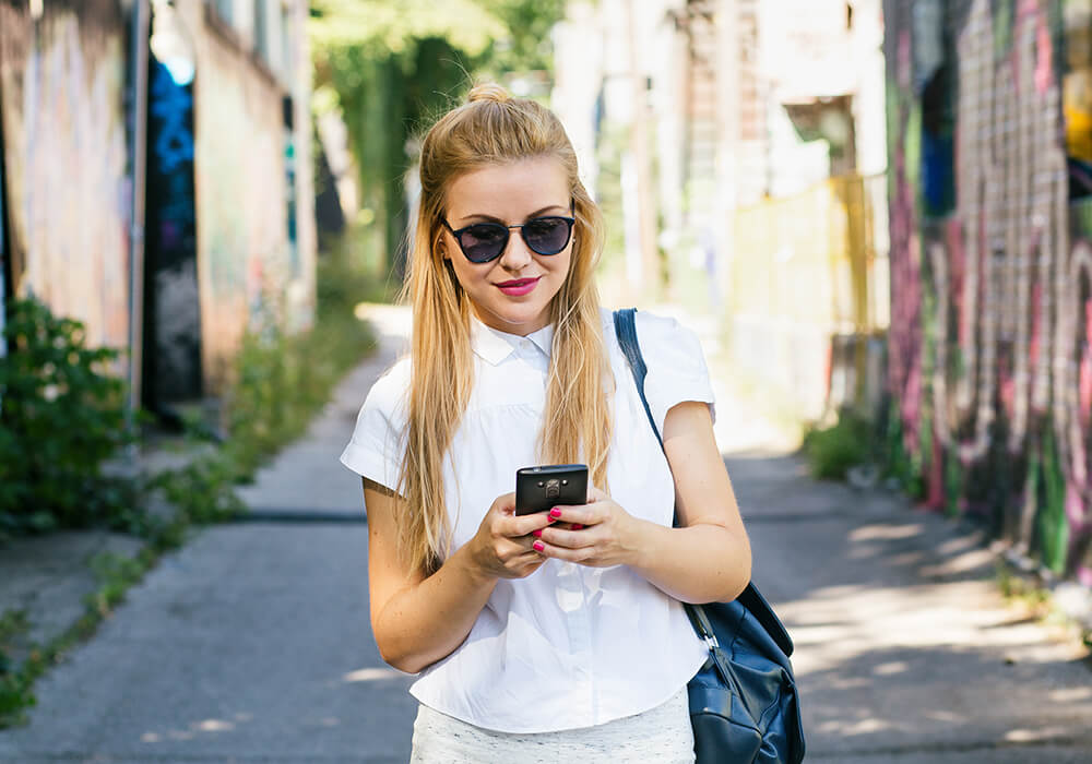 woman standing on sidewalk texting on phone wearing sunglasses