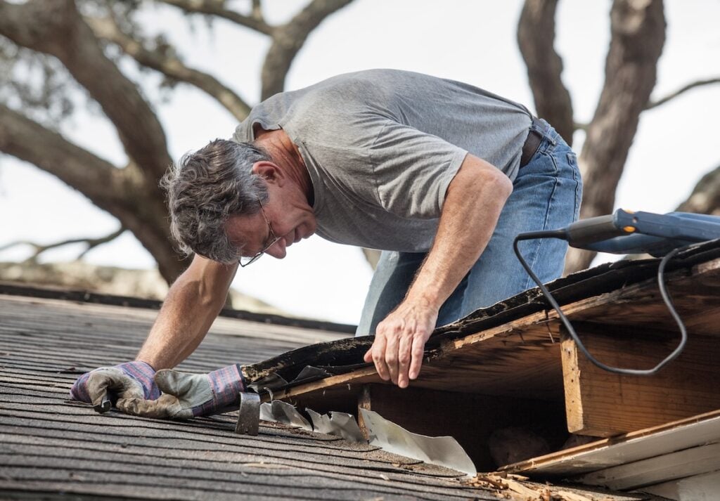 Close up view of man using crowbar and saw to remove rotten wood from leaky roof decking