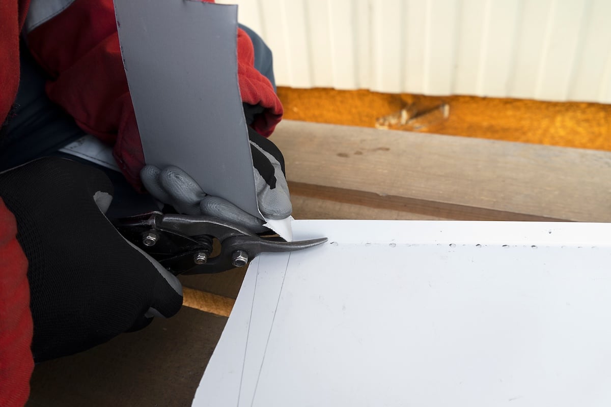 Builder with scissors cuts a white steel sheet. A working tinsmith cuts out an iron roof at a mark.