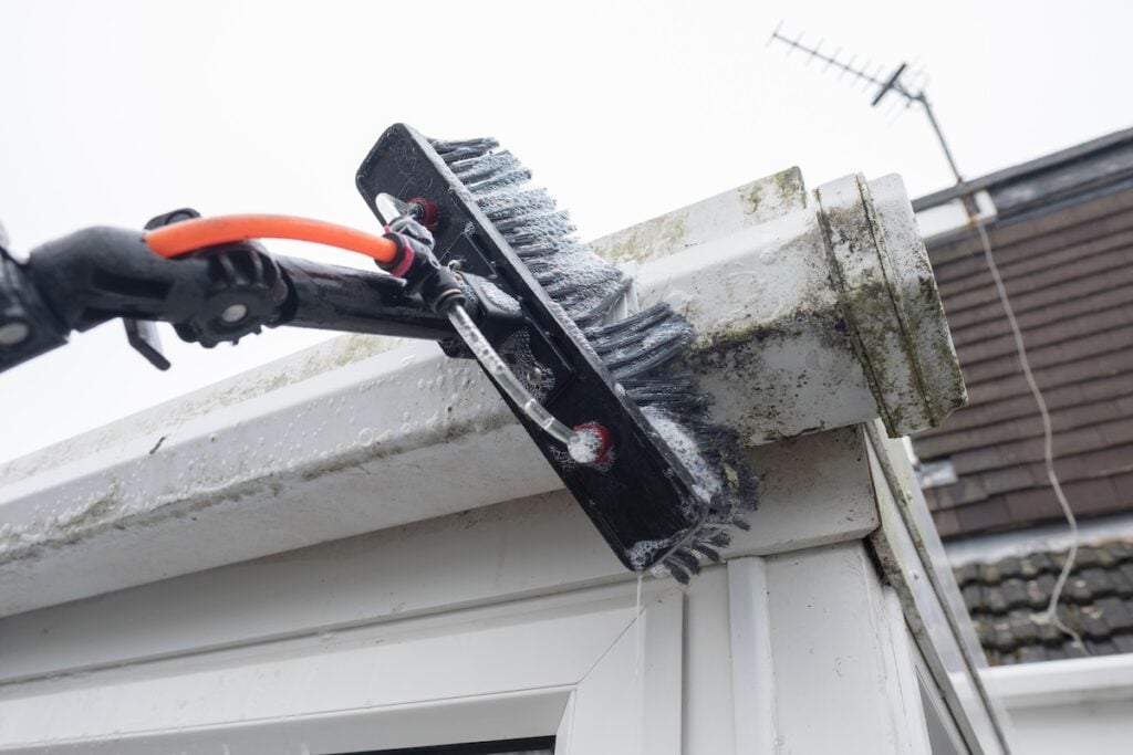A brush cleaning dirty clogged white plastic pvc gutters and drain pipes with mossy green mould plastic fascias.  